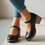Zwarte Mary Jane-schoenen met brogue-perforatie en bandje aan de voeten van een model in spijkerbroek, close-up op effen ondergrond 1