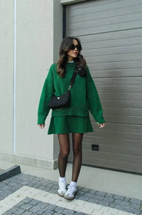Volledige look in groene gebreide set met zwarte panty, witte sneakers en zwarte schoudertas op een stoep. 9