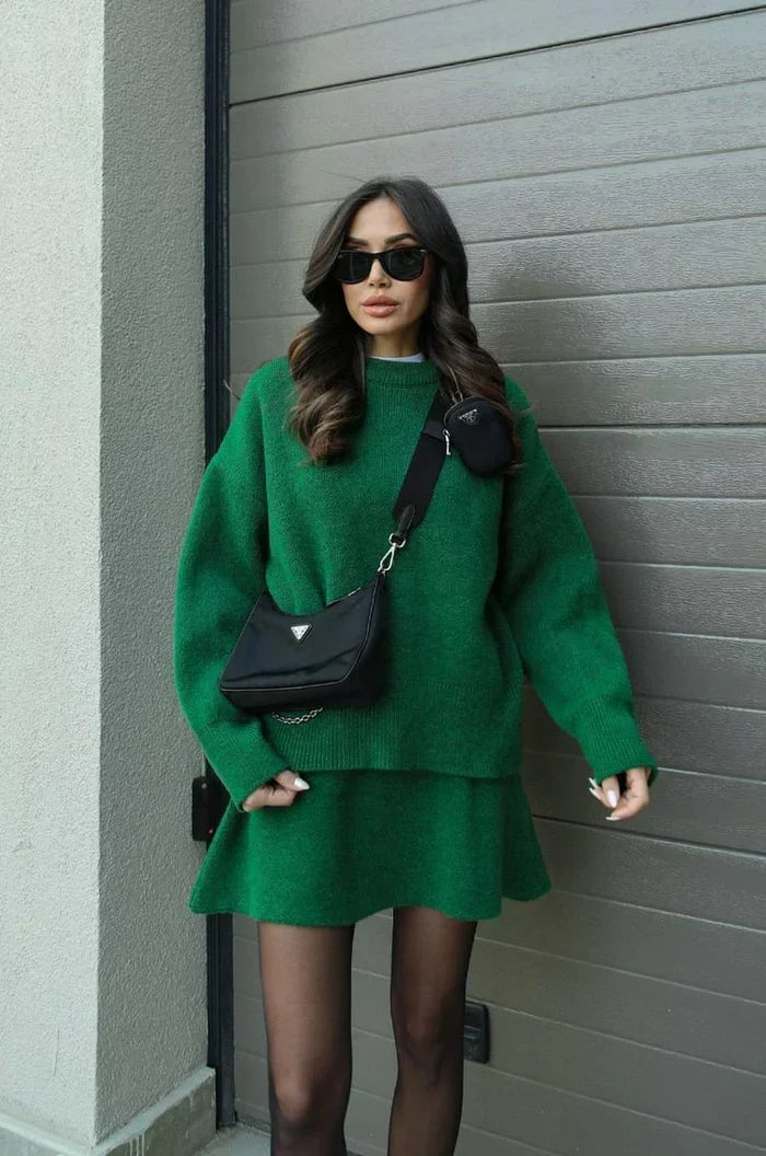 Model in groene gebreide trui en rok met zwarte schoudertas, zonnebril en zwarte panty voor een garagedeur. 8