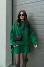 Model in groene gebreide trui en rok met zwarte schoudertas, zonnebril en zwarte panty voor een garagedeur. 8
