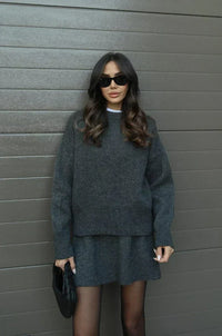 Model in antracietgrijze gebreide trui en rok met zwarte zonnebril en gevlochten zwarte clutch, frontaal. 11
