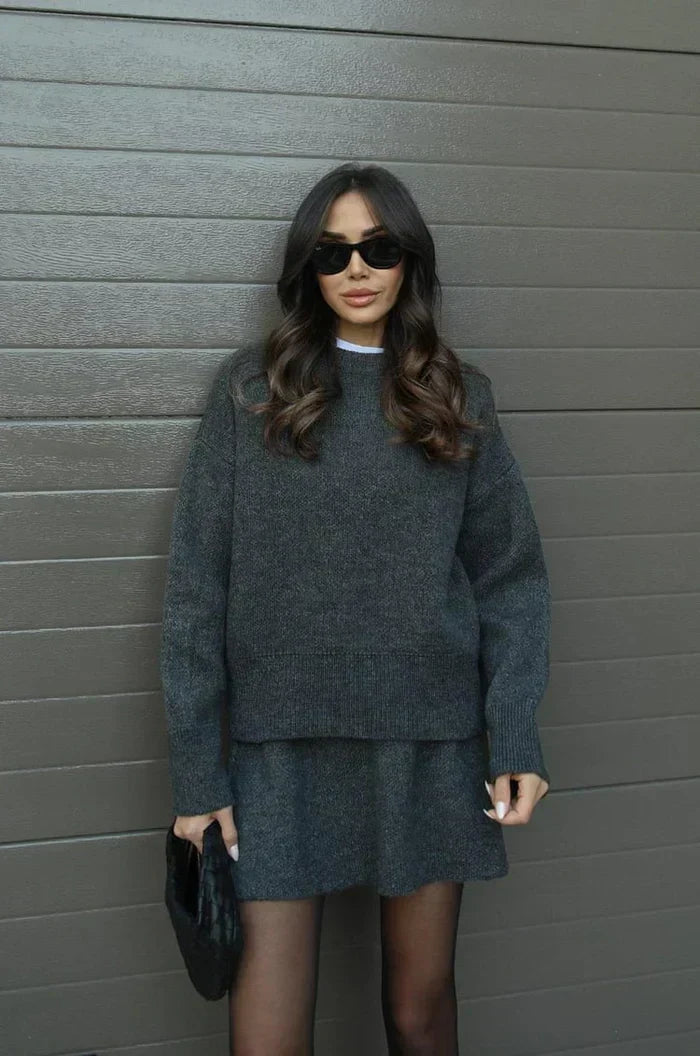 Model in antracietgrijze gebreide trui en rok met zwarte zonnebril en gevlochten zwarte clutch, frontaal. 11