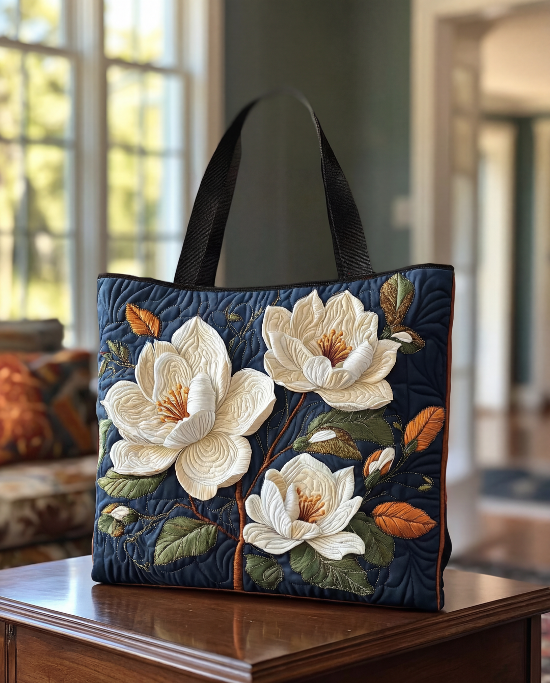Donkerblauwe gewatteerde draagtas met zwarte hengsels en grote witte magnoliabloemen met groene en oranje bladeren, op een houten tafel in een woonkamer. 1