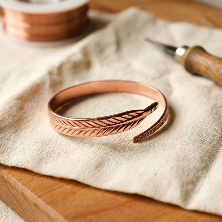 Open koperen armband met gegraveerd veermotief, liggend op een linnen doek naast een rol koperdraad en een graveergereedschap. 1