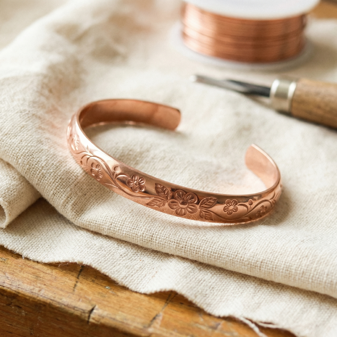 Open koperen armband met gegraveerde bloemen en ranken op linnen, naast graveergereedschap en een spoel koperdraad 1