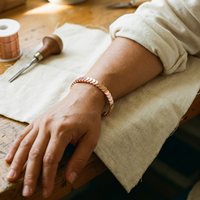 Koperen armband met overlappende schijven om een pols, rustend op een houten werktafel met linnen 2