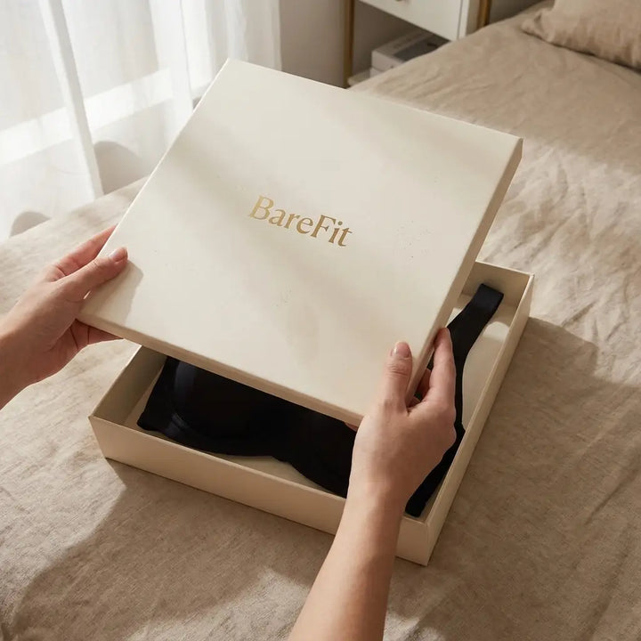 Handen openen een crèmekleurige geschenkdoos op een bed, waarin de zwarte bh netjes opgevouwen ligt. 14