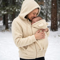 Vrouw in beige sherpahoodie kust haar baby, die in het paneel met beren-oortjes zit, in een besneeuwd bos. 8