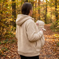 Vrouw van opzij in beige sherpahoodie draagt een baby in het babypaneel met capuchon op een herfstachtig bospad. 6