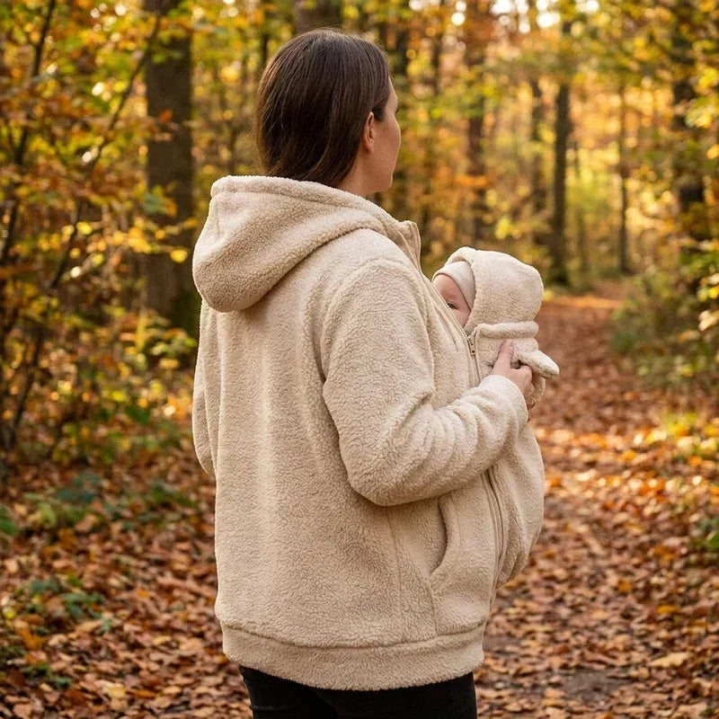 Vrouw van opzij in beige sherpahoodie draagt een baby in het babypaneel met capuchon op een herfstachtig bospad. 6