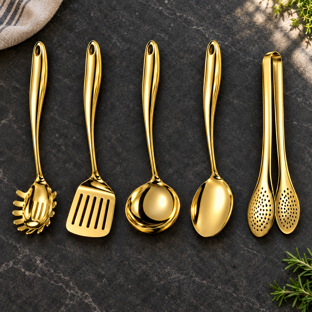 Vijfdelige set in glanzend goud op een donker stenen blad: pastalepel, spatel, soeplepel, lepel en tang, met theedoek en kruiden ernaast. 24