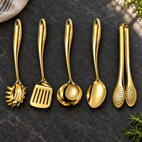 Vijfdelige set in glanzend goud op een donker stenen blad: pastalepel, spatel, soeplepel, lepel en tang, met theedoek en kruiden ernaast. 24