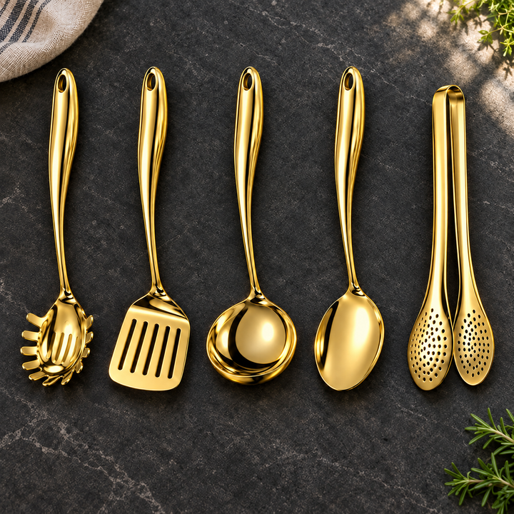 Vijfdelige set in glanzend goud op een donker stenen blad: pastalepel, spatel, soeplepel, lepel en tang, met theedoek en kruiden ernaast. 24