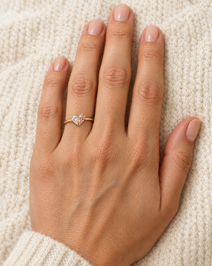 Hand met de gouden ring met helder en roze hartje om de ringvinger, tegen een crèmekleurige gebreide trui. 2