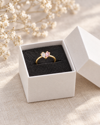 Gouden ring met helder en roze steen in een wit doosje met zwarte vulling, naast gedroogde witte bloemetjes. 3