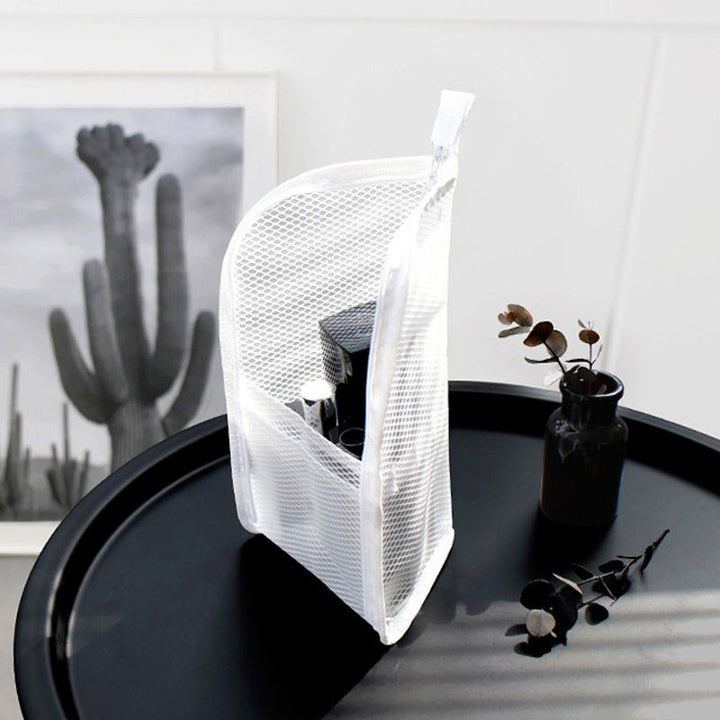 Witte mesh toilettas staat rechtop op een zwart tafeltje, gevuld met cosmetica, naast een vaasje met takjes en een cactusposter. 2
