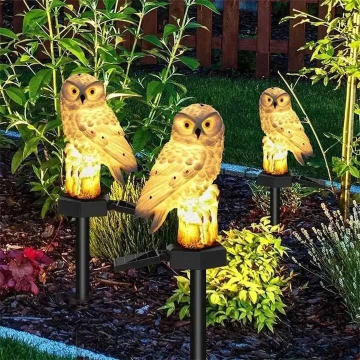Drie bruingele uilenlampen op palen, warm verlicht, tussen planten en bodembedekker in een tuinbed. 7