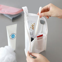 Hand haalt een tandenborstel uit de witte mesh toilettas, die overeind staat en gevuld is met tube, borstel en kleine spullen. 1