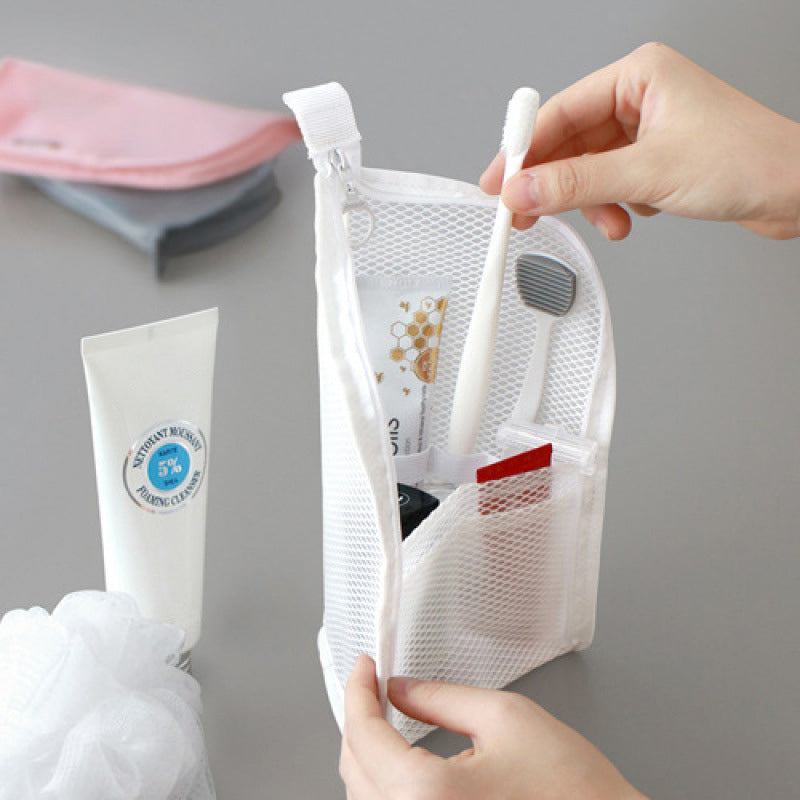 Hand haalt een tandenborstel uit de witte mesh toilettas, die overeind staat en gevuld is met tube, borstel en kleine spullen. 1