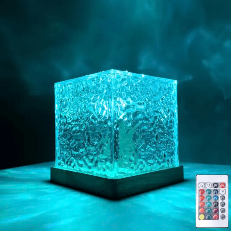 Doorzichtige kubuslamp met golvend oppervlak op houten voet, turquoise verlicht in het donker, met de afstandsbediening rechtsonder in beeld. 1