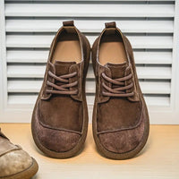 Paar donkerbruine suède veterschoenen met contrasterende stiksels, van voren gezien voor een wit jaloezieënpaneel. 10