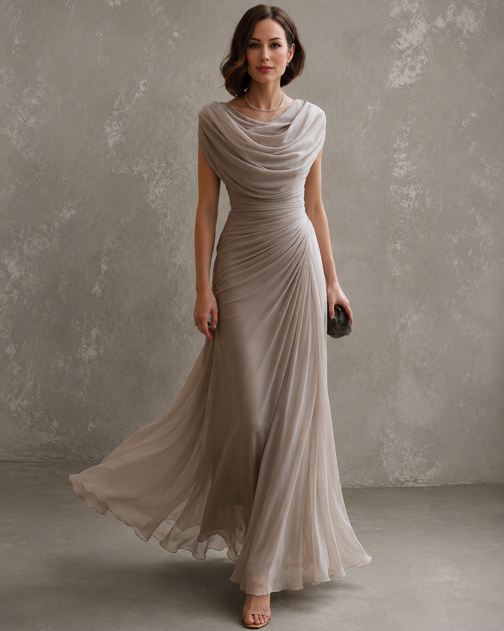 Model in taupegrijze maxi-jurk met watervalhals en gedrapeerde taille, de zwierige rok waait mee, met kleine clutch. 1