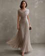 Model in taupegrijze maxi-jurk met watervalhals en gedrapeerde taille, de zwierige rok waait mee, met kleine clutch. 1