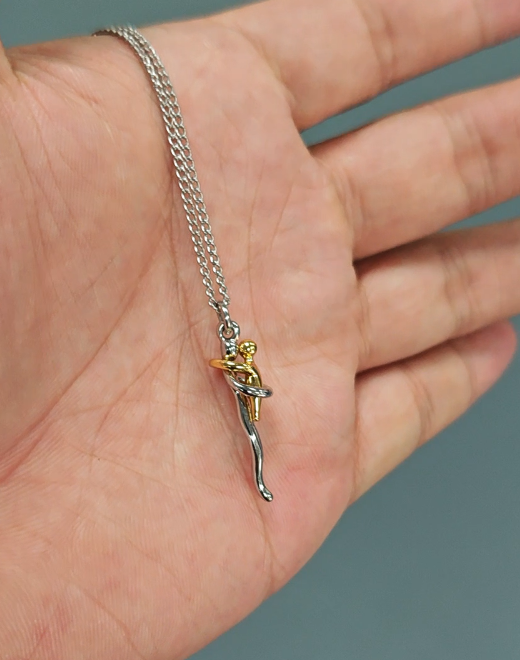 De ketting met zilver- en goudkleurige omhelzingshanger ligt op een open handpalm, wat het kleine formaat van de hanger laat zien. 7