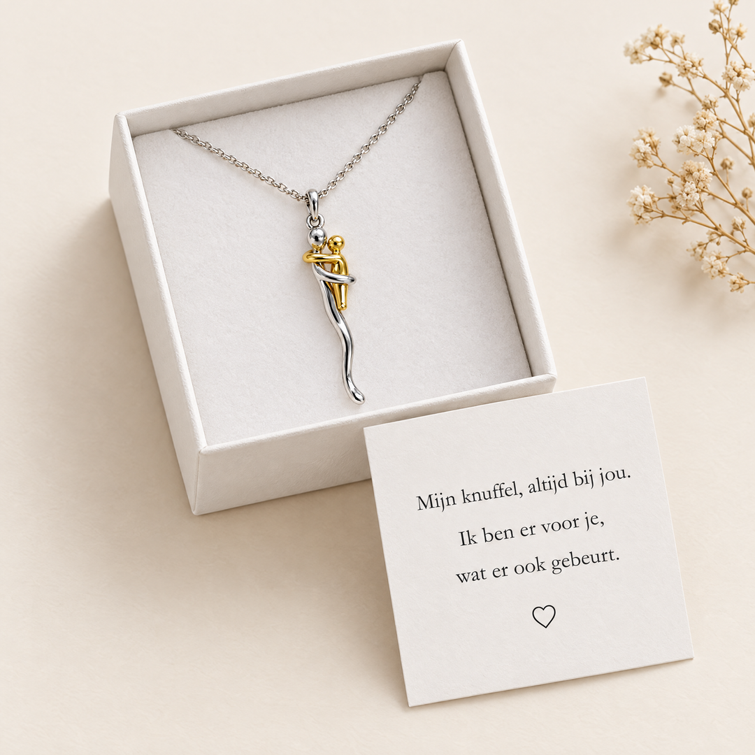 Ketting met omhelzingshanger in een open wit doosje, met een kaartje met een liefdevolle boodschap in het Nederlands ervoor en gedroogde bloemen ernaast. 2