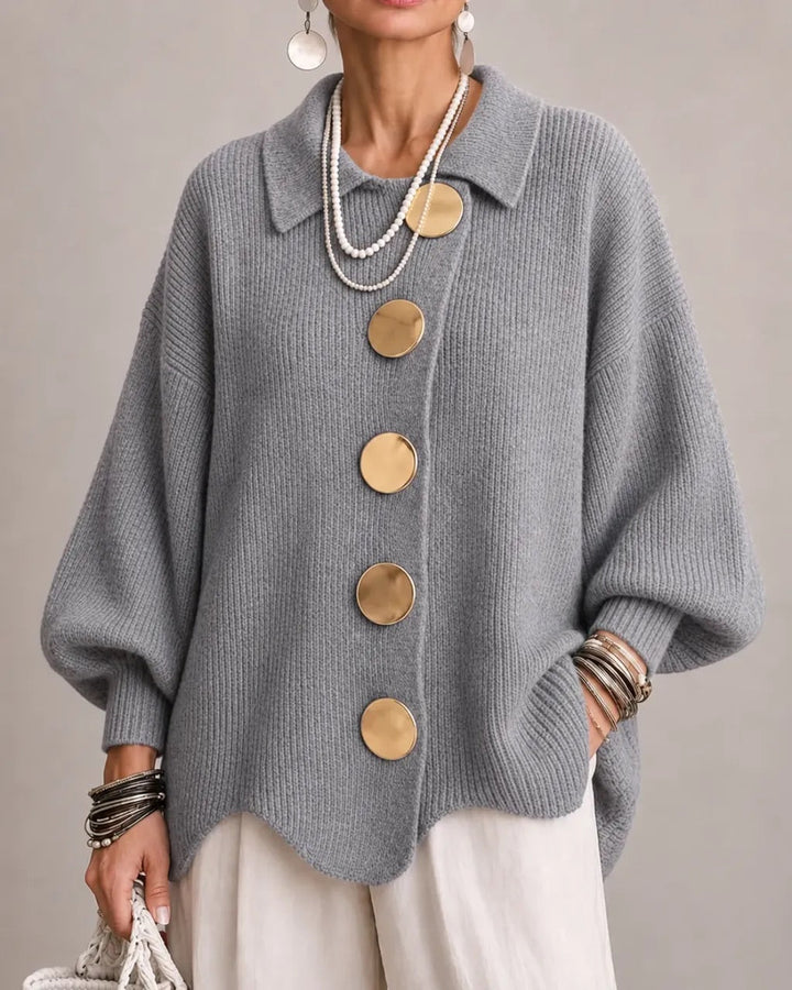 Model draagt het gebreide vest in grijs met gouden knopen, parelkettingen en een witte broek, vooraanzicht. 18