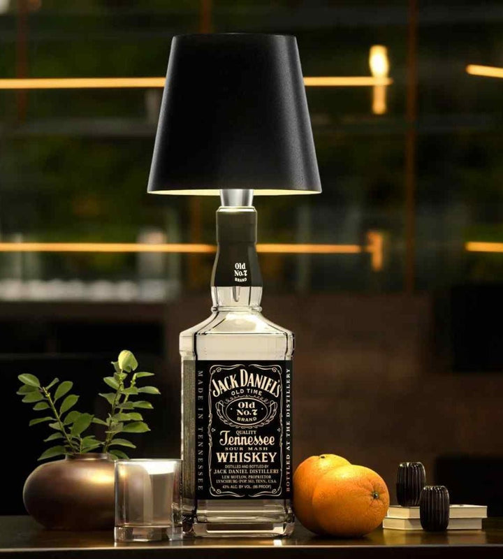 Zwarte lampenkap op een vierkante whiskyfles, met een glas, sinaasappels en een plant op een donkere bartafel. 2