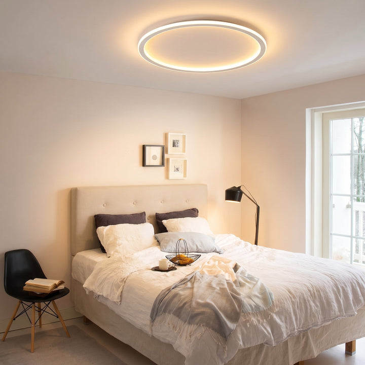 Lichtgevende ringlamp boven een slaapkamer met opgemaakt bed, gestoffeerd hoofdbord, zwarte vloerlamp en zwarte stoel met boek. 9