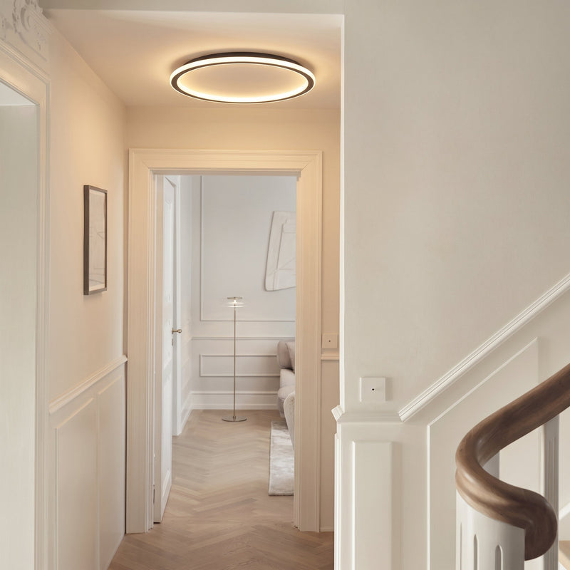 Lichtgevende ringlamp aan het plafond van een lichte gang met witte lijstwerk en een trapleuning van hout. 5