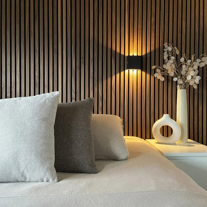 Zwarte wandlamp op houten lamellenwand naast een bed met kussens, vazen en gedroogde takken 4
