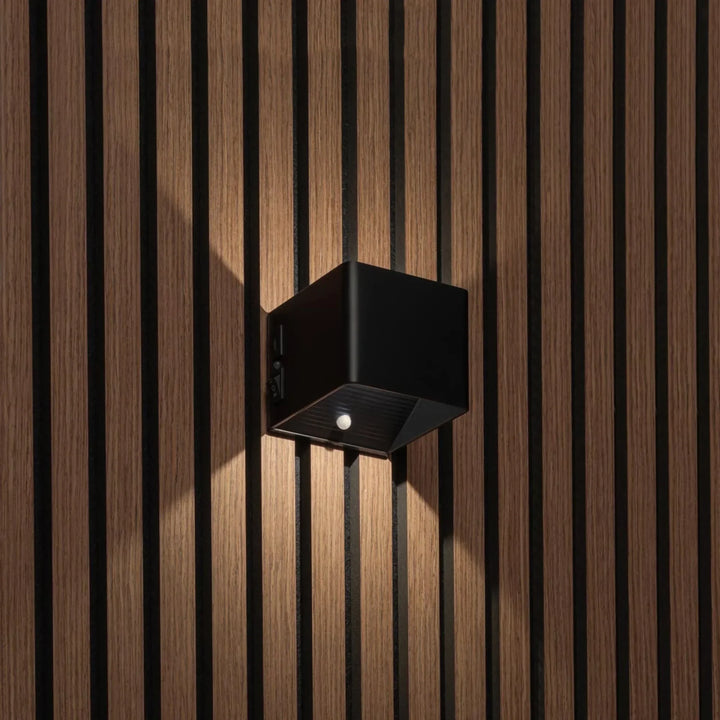 Zwarte kubusvormige wandlamp op houten lamellenwand, schuin gezien met bewegingssensor, schakelaar en lichtstralen omhoog en 1