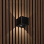 Zwarte kubusvormige wandlamp op houten lamellenwand, schuin gezien met bewegingssensor, schakelaar en lichtstralen omhoog en 1