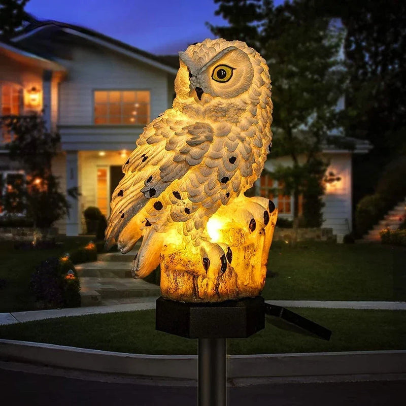 Witte uil met zwarte stippen en warm lichtende boomstronk op een paal, 's avonds voor een verlicht huis. 2