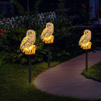 Drie uilenlampen op palen met warm licht langs een tuinpad in het donker, tussen struiken en planten. 3