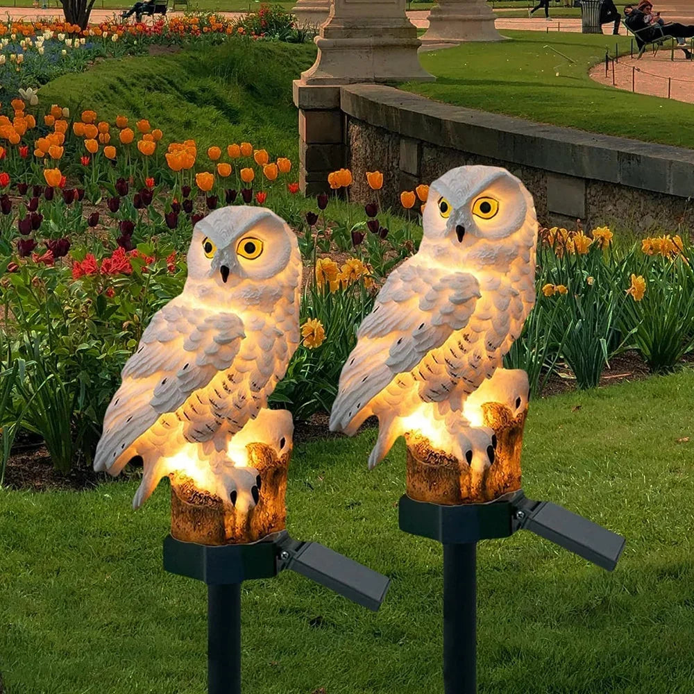 Twee wit verlichte uilenlampen op een paal in een tuin met oranje en gele tulpen en narcissen. 1
