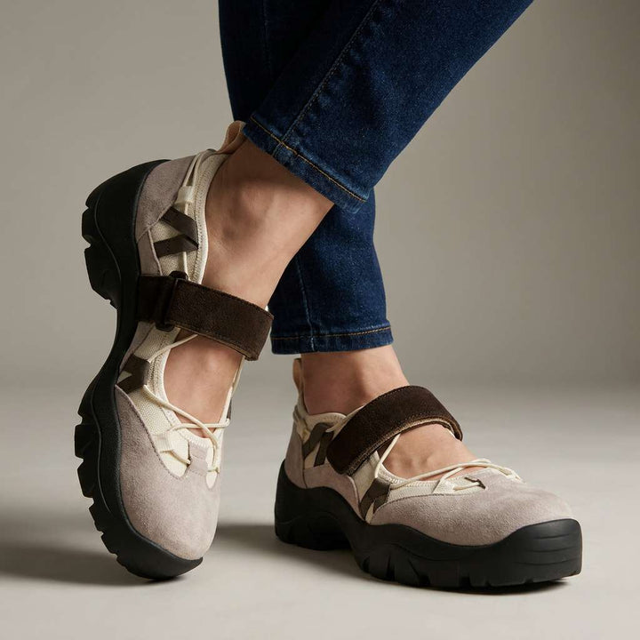 Beige suÃ¨de instapschoenen met bruine klittenbandband en dikke zwarte profielzool, gedragen met donkere jeans tegen een neutrale studioachtergrond. 1