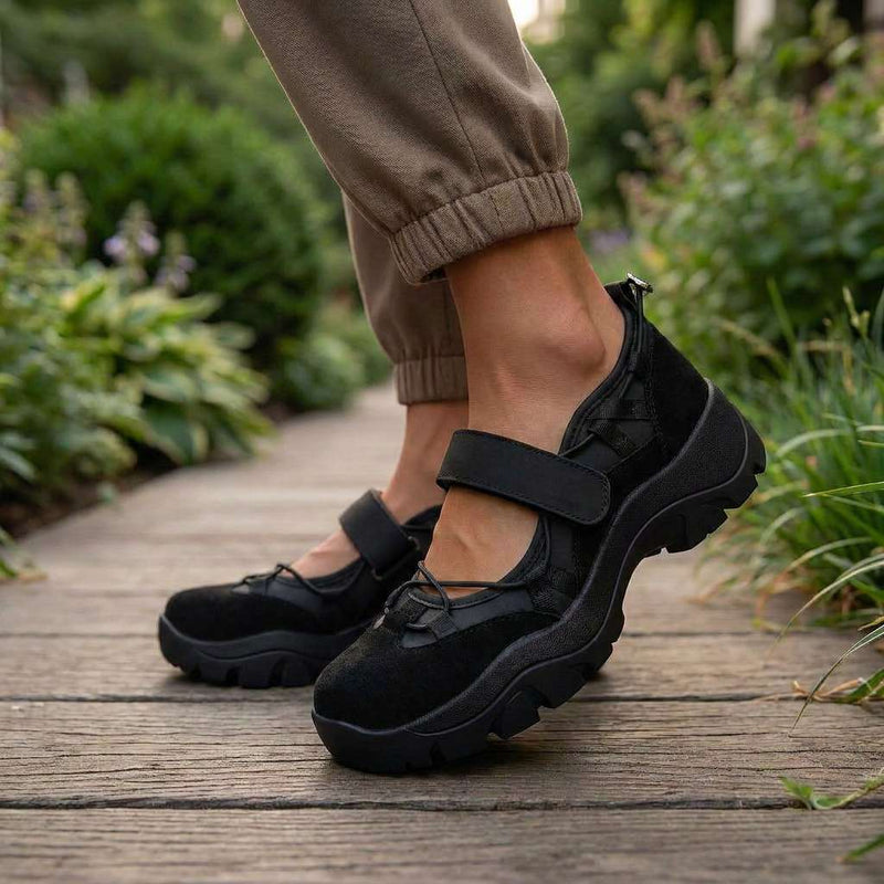 Zwarte schoenen met klittenbandband, gedragen met bruine broek op een houten pad omringd door tuinplanten. 10