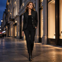 Vrouw loopt 's avonds door een winkelstraat in een zwarte structuurblazer met zwarte top, pantalon en enkellaarsjes. 3