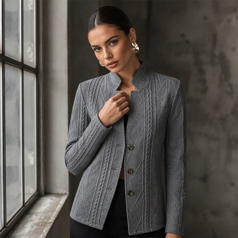 Vrouw bij een raam in een grijze blazer met kabelpatroon; ze houdt de opstaande kraag met één hand vast. 10