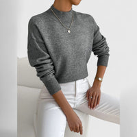 Model draagt de grijsgemêleerde trui met ribkraag en brede ribboorden, met witte broek en een dunne ketting met hanger, zittend op een witte fauteuil. 12