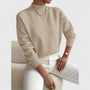 Model draagt de trui in beige met ribkraag en ribboorden, gecombineerd met witte broek, gouden kettingen en horloge. 7