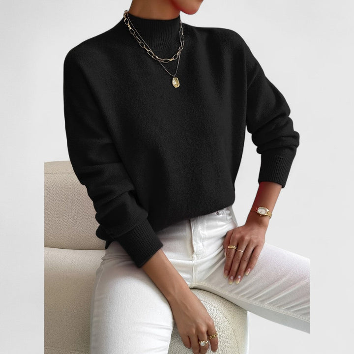 Model draagt de zwarte trui met ribkraag en ribboorden, met witte broek en gouden sieraden, zittend op een fauteuil. 8