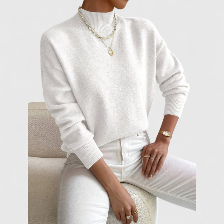 Model draagt de trui in gebroken wit met witte broek, gouden kettingen en gouden horloge, zittend op een bouclé fauteuil. 4