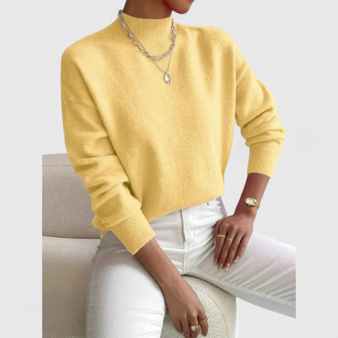 Model draagt de trui in zachtgeel met ribkraag en ribboorden, met witte broek en een bouclé fauteuil op de achtergrond. 6