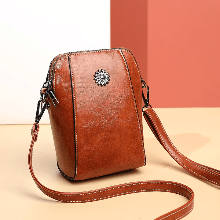 Roodbruine leren crossbodytas schuin van voren, met bloemvormig sierstuk en lange band op beige ondergrond 1