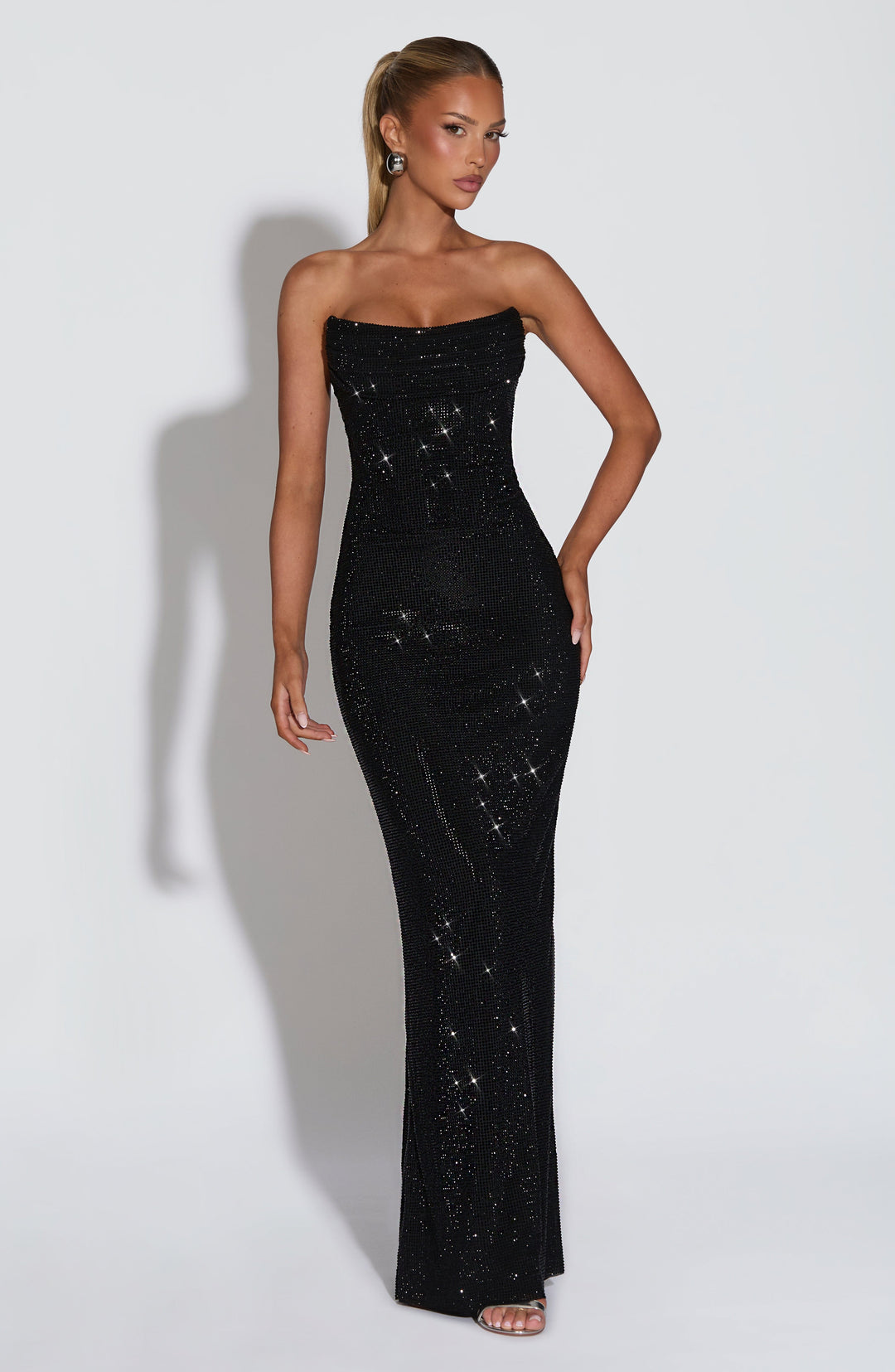 Vooraanzicht van de zwarte strapless maxi-jurk met strass op een model, hand op de heup, strakke pasvorm tot op de vloer. 22